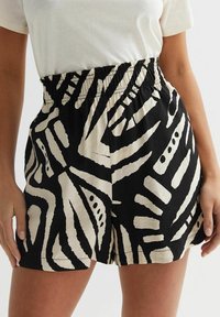 Pantaloni corti neri e crema con una fascia elastica in vita. Presentano linee e forme astratte audaci sullo sfondo nero.