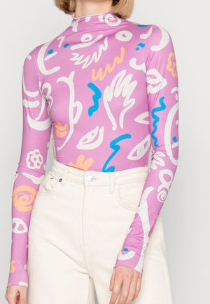 Femme portant un haut moulant à manches longues lilas avec des motifs abstraits blancs, bleus et pêches, associé à un pantalon crème taille haute.