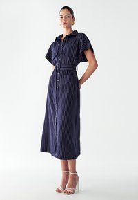 Tumestriipidega nööbitav kleit lühikeste varrukatega, vöö taljes ja kaks küljetaskut. Kombineeritud valgete kontsakingadega.