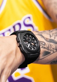 Black analog wristwatch with octagonal case, textured strap, multi-function dial with orange accent, worn on tattooed wrist.