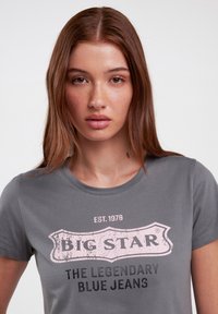 Pilkas medvilninis marškinėliai su apvali kaklo iškirpte ir trumpomis rankovėmis. Priekinis piešinys apima užrašą "BIG STAR" rožine spalva ir "THE LEGENDARY BLUE JEANS" juoda spalva.