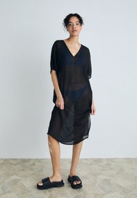 Black sheer cover-up dress with a V-neck, short sleeves, and a loose fit, paired with black slide sandals on a textured surface.