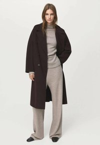 Long manteau brun foncé sur un pull gris clair et un pantalon gris à jambes larges. Textures lisses et coupe décontractée, avec des chaussures noires.