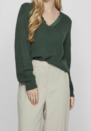 Femme portant un pull en maille vert foncé par-dessus un haut vert bordé de dentelle, associé à un pantalon beige taille haute sur un fond uni.