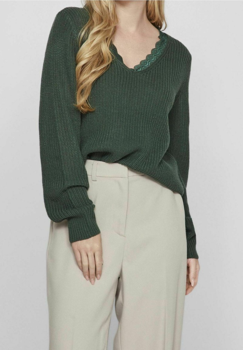 Femme portant un pull en maille vert foncé par-dessus un haut vert bordé de dentelle, associé à un pantalon beige taille haute sur un fond uni.