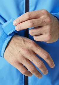 Lichtblauwe jas met een klittenband manchette. Een hand past de manchette aan en onthult een gladde textuur en een rits in het midden. Vlekkerige huid zichtbaar.