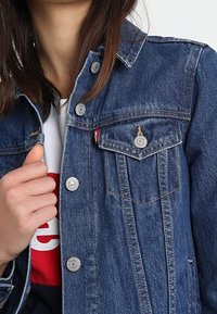 Person wearing a blue denim jacket buttoned at the top over a white and red graphic shirt, holding the jacket near the collar.