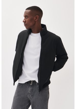 Zwarte zip-up jasje met een staande kraag, gedragen over een wit T-shirt en grijze jeans. Het jasje heeft elastische manchetten en een strak ontwerp.