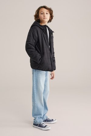 Mlada oseba stoji, oblečena v črno puhasto jakno, svetlo modre široke kavbojke in mornarsko modre superge, na nevtralnem ozadju.