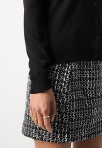 Svart stickad cardigan med en texturerad fåll, tillsammans med en högmidjad svart och vit tweedkjol. Guldringar pryder fingrarna.