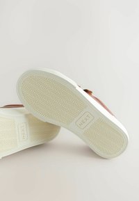 Hnědé semišové tenisky s bílými gumovými podrážkami, které mají diagonální vzor a označenou plochu. Design je ležérní a nízkoprofilový.