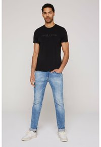 Camiseta de algodón negra con un logo texturizado, combinada con unos vaqueros de mezclilla azul claro. El calzado son zapatillas blancas. Diseño simple y casual.