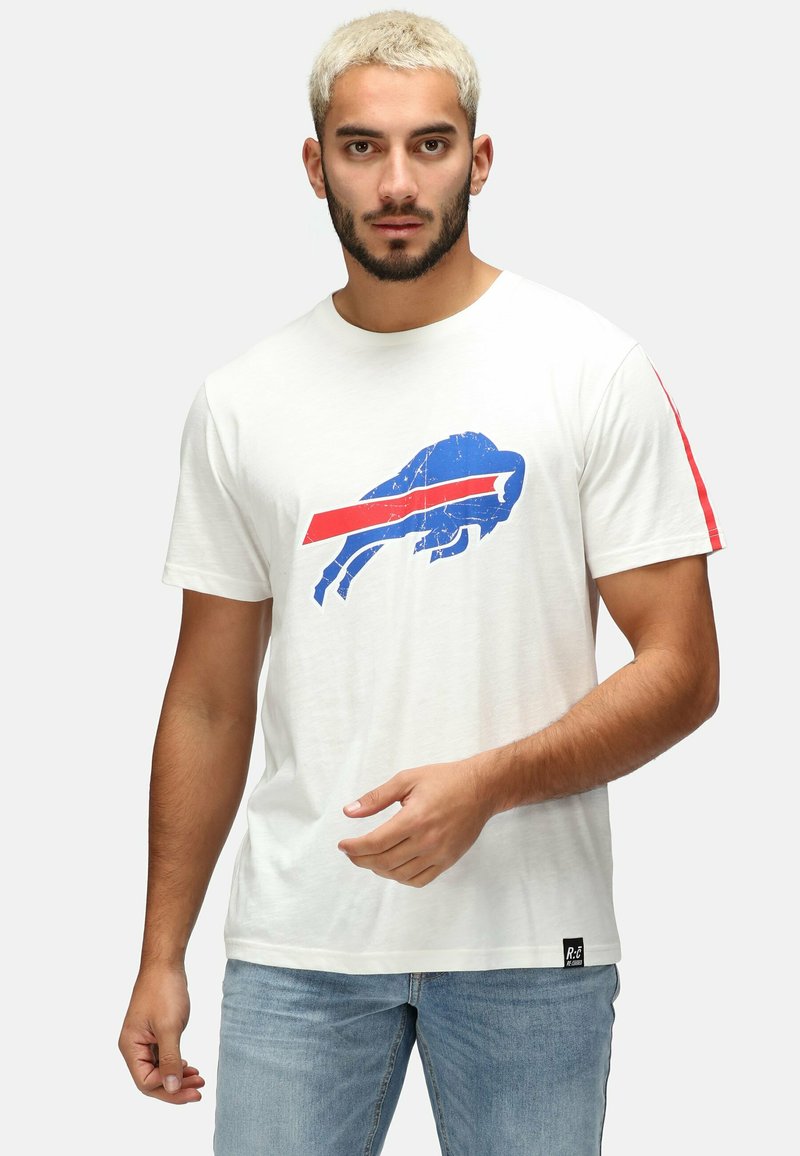 T-shirt en coton blanc avec un logo Buffalo bleu et rouge. Présente des rayures rouges sur les manches et une étiquette à l'ourlet. Porté avec un jean bleu clair.