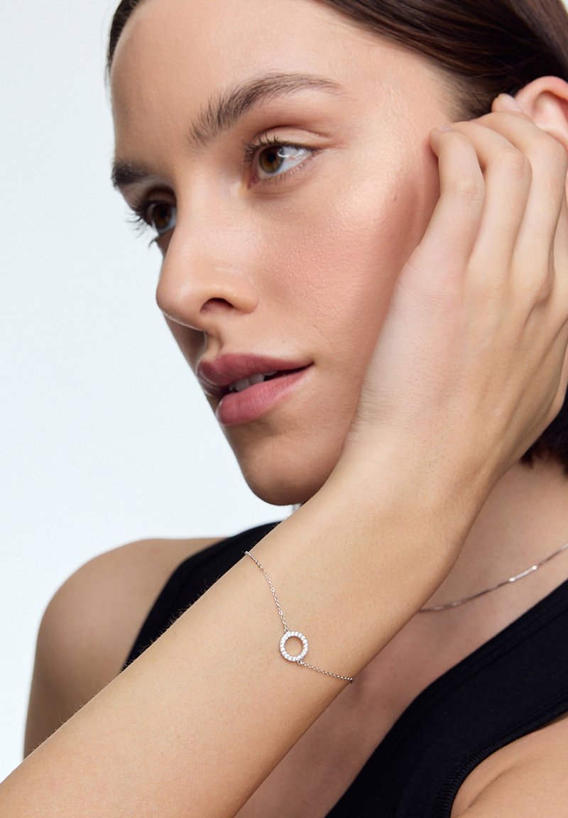 Srebrna bransoletka z okrągłym wisiorkiem, ozdobiona małymi przezroczystymi kamieniami wokół krawędzi. Nadgarstek modelki jest częściowo widoczny na neutralnym tle.