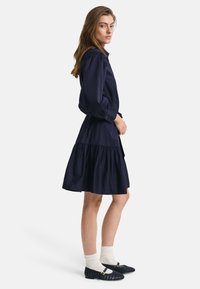 Rochie din bumbac bleu marin cu deschidere cu nasturi în față, mâneci lungi tip balon și tiv cu volane în straturi. Poartă pantofi plată țesute negri și șosete albe.