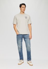 Beige katoenen t-shirt met print op de linkerborst, gecombineerd met blauwe denim jeans en lichtgekleurde sneakers. Casual fit, korte mouwen.