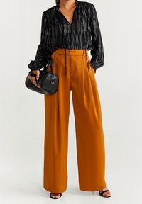 Mujer con blusa negra estampada, pantalones de pierna ancha mostaza de cintura alta, sosteniendo un bolso negro y usando tacones abiertos negros.