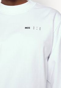 Sweat-shirt blanc à col rond porté par une personne à la peau foncée, avec un petit texte noir "MCQ" et un motif abstrait sur le côté gauche de la poitrine.