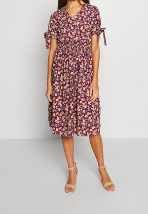 Kobieta ubrana w bordową sukienkę do kolan w kwiatowy wzór z falbankami na rękawach zawiązanymi na kokardki oraz beżowe sandały na kostkę, stojąca na szarej podłodze.