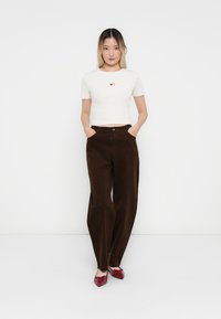 Witte geribde crop top met een rood hartprint, gecombineerd met bruine wijde corduroy broeken en glanzende rode schoenen, model staat met handen in de zakken.