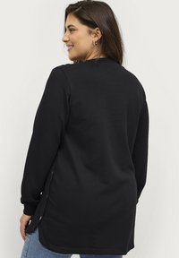 Camiseta negra de manga larga con cuello redondo, botones laterales y dobladillo curvado, combinada con jeans de denim azul. Textura de tela suave.