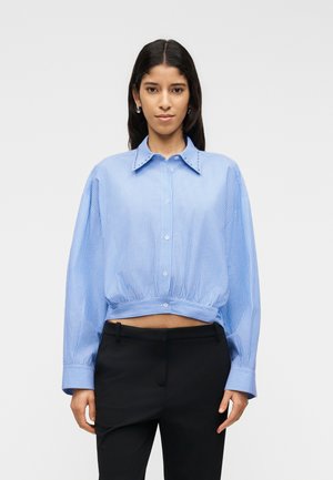 Žena nosi modro-bielu pruhovanú krátku košeľu s gombíkmi a prehnanými rukávmi a čierne nohavice, stojaca pred jednoduchým svetlým pozadím.