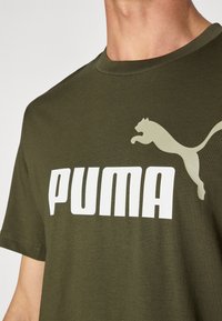 Olajzöld pamut póló kerek nyakkal, fehér "PUMA" logóval és világoszöld puma sziluettel a bal felső részen.