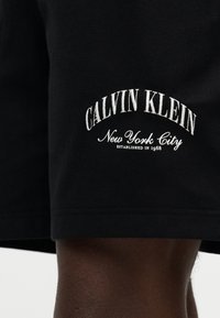 Μαύρο ύφασμα με λευκό κεντημένο κείμενο "Calvin Klein New York City Established in 1968" πάνω από το γόνατο ενός ατόμου με σκουρόχρωμο δέρμα.