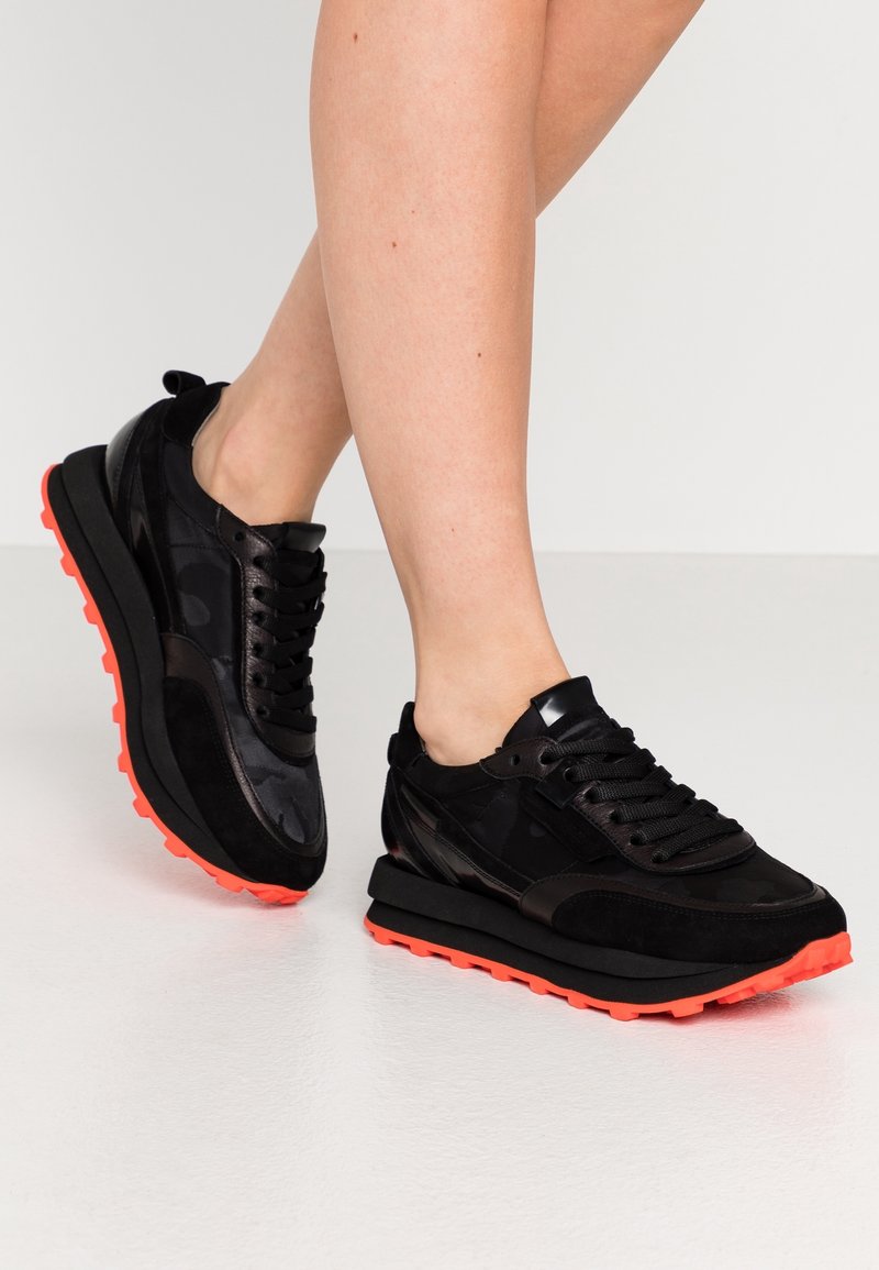 Jambes portant des baskets noires avec des semelles orange vif, debout sur un sol blanc lisse.