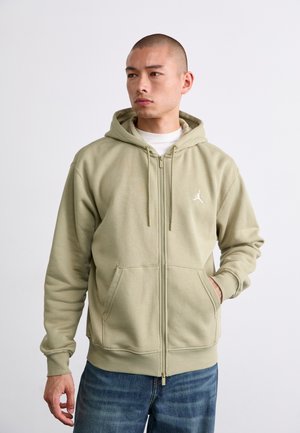 Light green zip-up hoodie made of soft fabric, featuring a front kangaroo pocket and a small white logo on the chest. Paired with blue jeans.