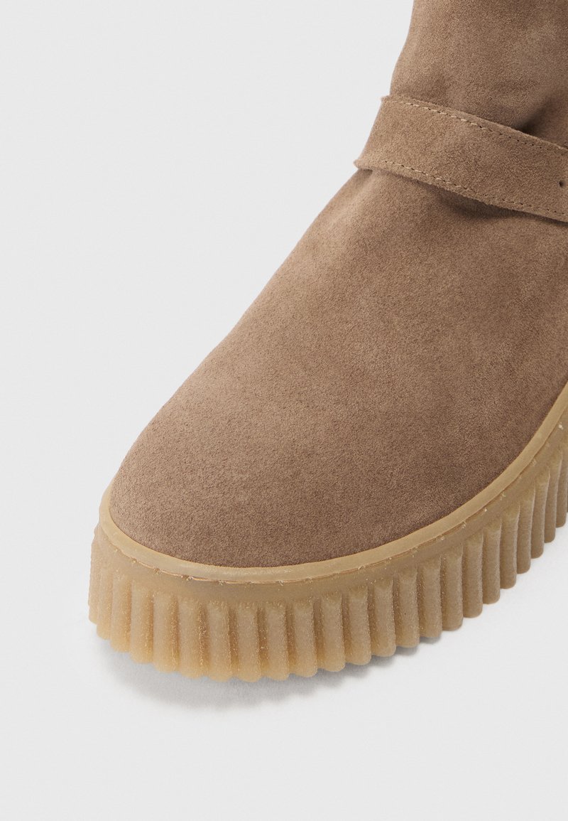 Botín de ante marrón con una suela beige gruesa y estriada, y un detalle de correa. Textura suave y forma de puntera redonda.