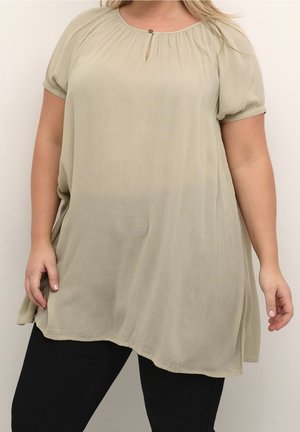 Tunique ample beige clair à manches courtes avec un petit détail bouton œillet à l'avant, portée avec un pantalon noir.