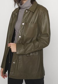 Olivgrüne Lederjacke mit einem kragenförmigen Ausschnitt, Druckknöpfen, zwei Fronttaschen und einer lockeren Passform. Weiche Textur mit glatter Oberfläche.