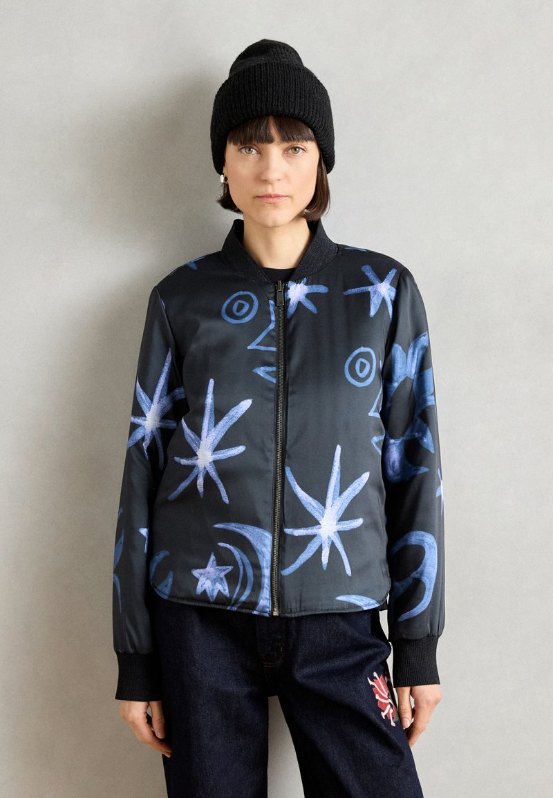 Veste bomber noire avec des motifs abstraits bleus en étoiles et tourbillons, col et poignets côtelés. Associée à un jean foncé et un beanie en tricot noir.