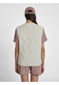 Quilted vest in light beige, featuring a rounded design and textured pattern. Paired with a pink short-sleeve shirt and shorts.