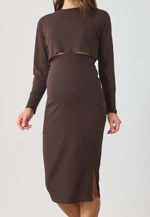 Femme portant une robe marron à manches longues en maille côtelée, avec une fente et une découpe au-dessus de la taille.