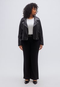 Μαύρο δερμάτινο μπουφάν μοτοσικλέτας με φερμουάρ, φορεμένο πάνω από άσπρο τοπ και μαύρο ψηλόμεσο παντελόνι. Το μοντέλο στέκεται με τα χέρια στα πλάγια.