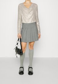 Metallic stříbrný dlouhý rukáv top s volným výstřihem, šedá kostkovaná plisovaná sukně, šedé punčochové ponožky do kolen a černá taška s kulatými kovovými detaily.