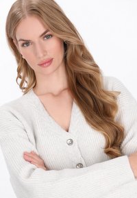 Világosszürke, bordázott kötött kardigán mély V-nyakkal és három nagy ezüstgombbal. Hosszú, kényelmes ujjak és puha textúra jellemzi.