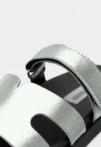 Silberne metallische Sandale mit einem breiten Zehenriemen mit Ausschnittdesign, schmalem Knöchelriemen, schwarzer Sohle und strukturiertem Finish.