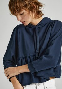 Sudadera con capucha azul marino, con cintura fruncida, mangas largas y un cordón texturizado. Tejido suave, diseño corto, con detalles mínimos.