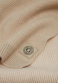 Tejido de punto acanalado de color crema con una textura suave, que presenta una etiqueta marrón con el logo en la esquina inferior, de la marca "Guess."