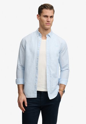 Mężczyzna z krótkimi włosami, ubrany w jasnoniebieską koszulę na guziki na białą koszulkę, czarne spodnie i zegarek na rękę, stojący z jedną ręką w kieszeni.