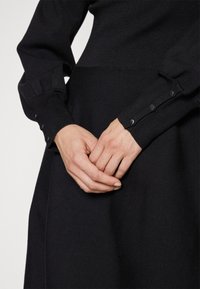 Les mains d'une femme jointes sur une robe noire à manches longues avec des poignets boutonnés, vues de la taille jusqu'au milieu des cuisses sur un fond neutre.