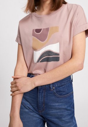 Osoba ubrana w bluzkę o krótkim rękawie w kolorze mauve z abstrakcyjnym nadrukiem artystycznym oraz wysokim stanem niebieskie dżinsy, stojąca z rękami skrzyżowanymi na wysokości talii.