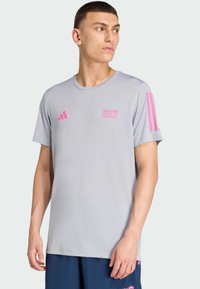 Mladić u svijetlosivoj BMW Berlin Marathon majici s ružičastim logotipima i tamnoplavim šortsem, gleda ulijevo na jednobojnoj pozadini.