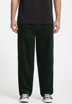 Corduroy pants in dark green, featuring wide legs and vertical ribbed texture. Paired with black sneakers featuring white laces and soles.
