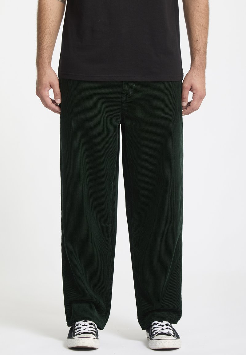 Donkergroene corduroy broek met wijde pijpen en verticale ribbelstructuur. Gepaard met zwarte sneakers met witte veters en zolen.