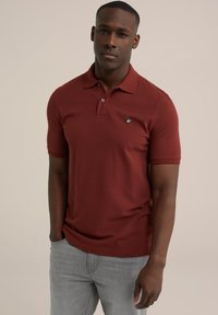 Polo de culoare maro cu mâneci scurte, cu guler cu două butoane și un mic logo brodat. Asortat cu blugi gri deschis, evidențiind o croială casual.