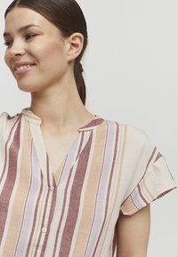 Blouse textile rayée avec un décolleté en V, des manches courtes et des détails de boutons, dans des teintes de beige, lavande et bordeaux avec une texture légère.